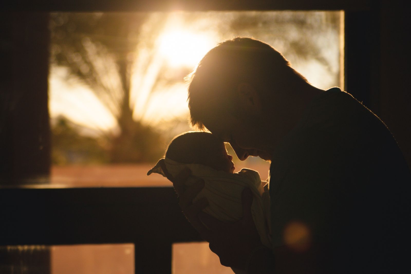 A father cradling his baby, their foreheads touching, silhouetted against a warm sunset
